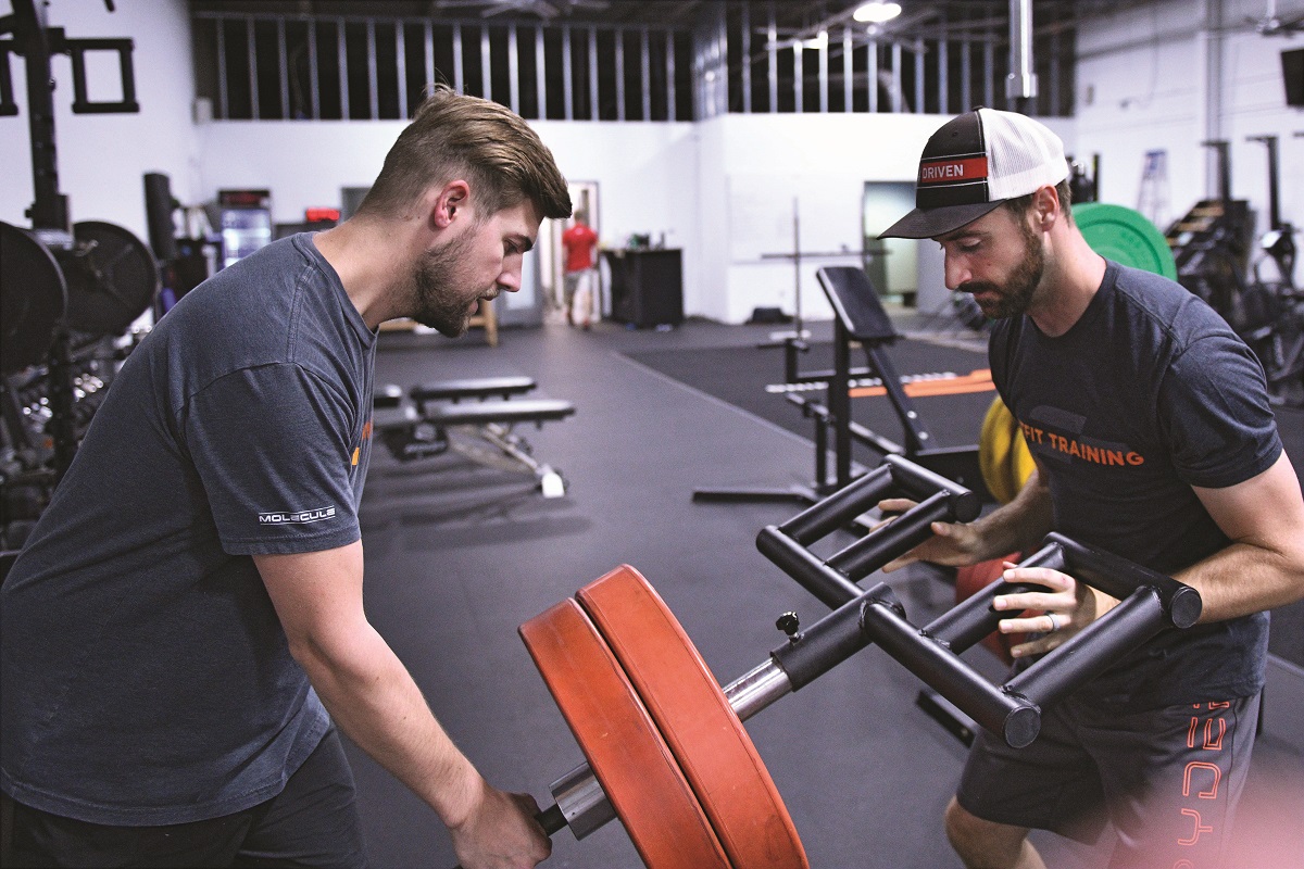 Two men in t-shirts stand on either side of a fitness machine in a gym