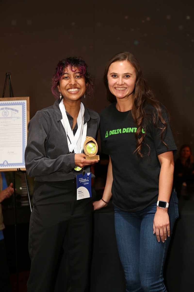 A girl in a black shirt and top holds an award and smiles for the camera next to a woman in a black t-shirt and jeans
